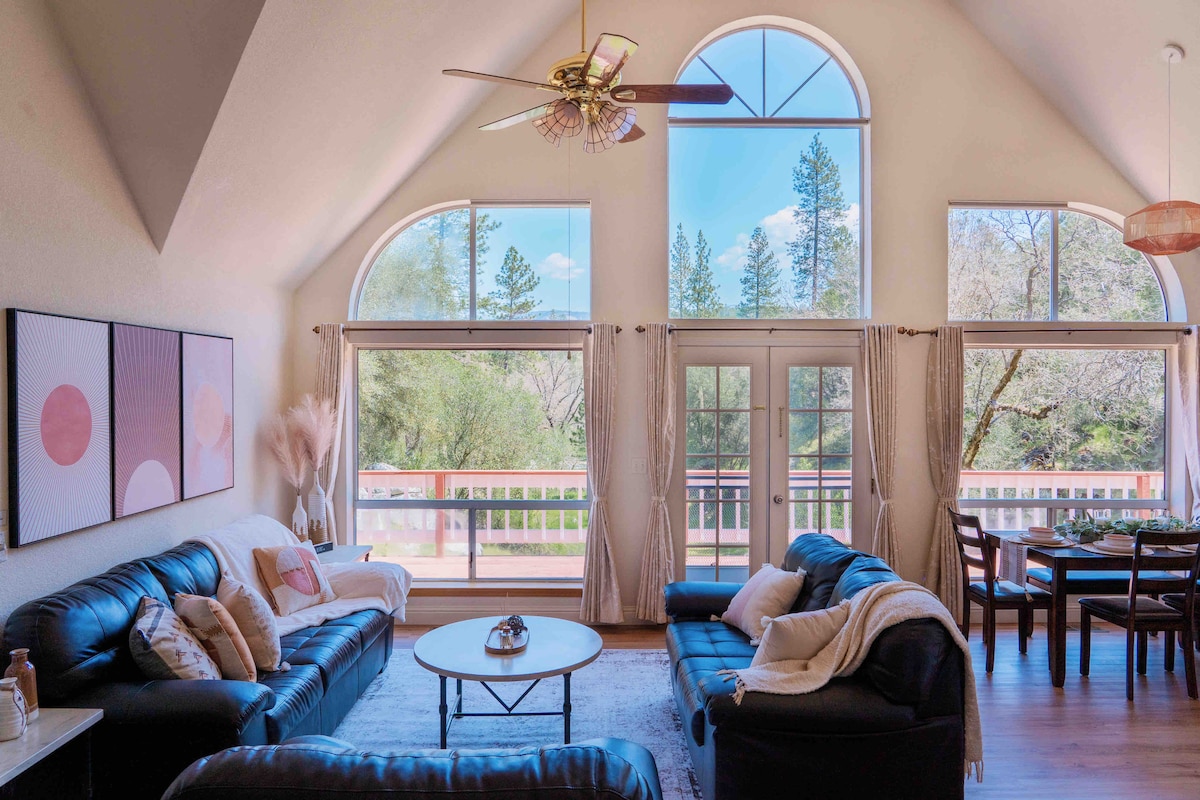 The spacious living area features large arched windows that invite natural light and offer views of the surrounding trees. Comfortable leather sofas surround a circular coffee table, creating a relaxing space. A dining area with a wooden table is seen in the background, merging indoor and outdoor elements.