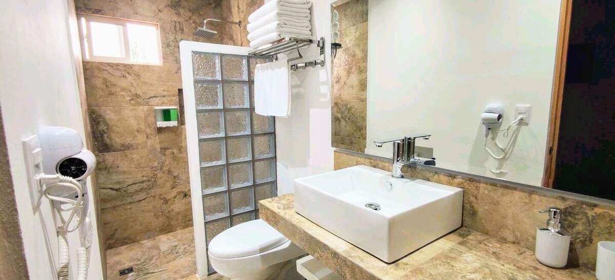 A modern bathroom features a glass shower enclosure and a spacious countertop with a square sink. Clean towels are neatly arranged above the sink. Natural light filters in through a small window, illuminating the textured stone walls and stylish fixtures.