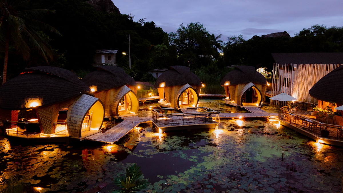 Several uniquely designed accommodations are situated around a serene pond featuring water lilies. Soft lighting highlights the curved architecture and wooden walkways that connect the units. Lush greenery frames the scene, creating a tranquil atmosphere in the evening light.
