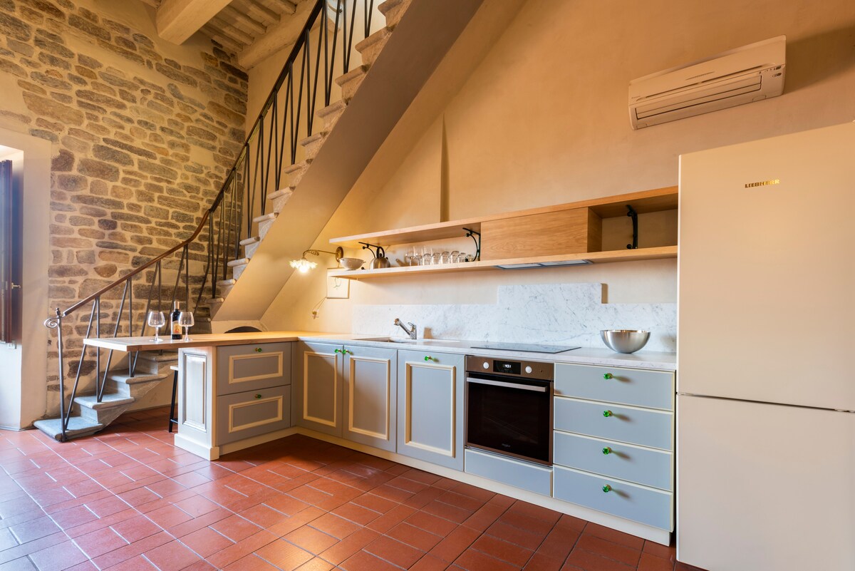 A well-equipped kitchen features light-colored cabinetry with green handles and a spacious countertop. Modern appliances include an oven and a refrigerator. Natural light enters from a nearby window, complementing the exposed stone wall and rustic staircase leading to the upper level.