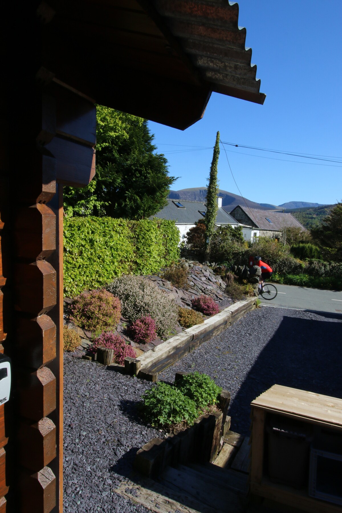The image captures a view from a cabin entrance, featuring a well-maintained garden with colorful shrubbery and a gravel pathway. In the foreground, a cyclist is seen riding along the quiet street, framed by greenery and distant hills under a clear blue sky.