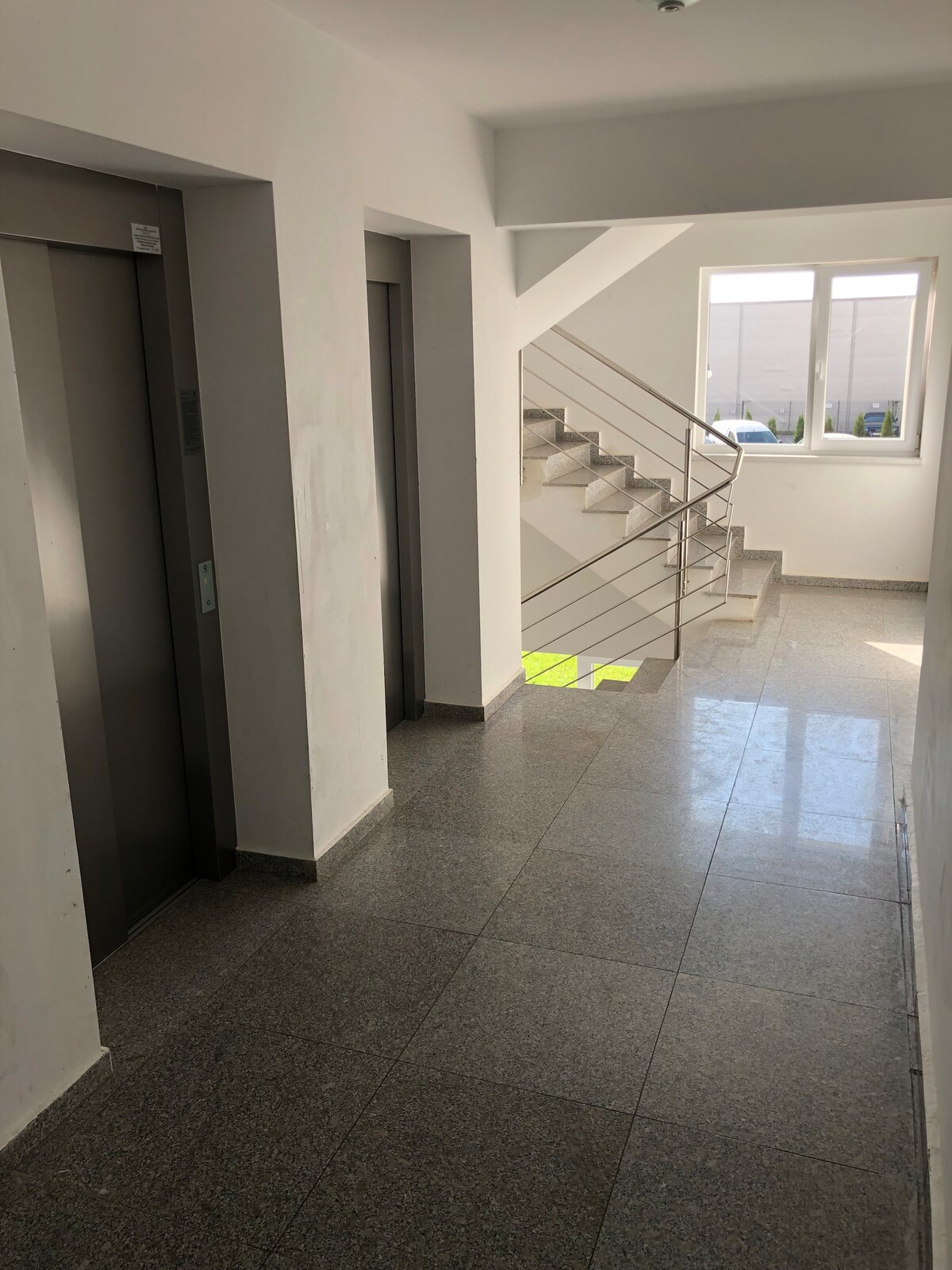 A bright corridor features two elevators and a staircase with modern metal railings. Natural light enters through a large window, illuminating the polished floor. The walls are painted white, enhancing the spacious feel of the area.