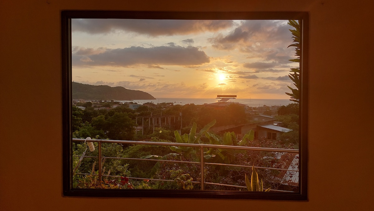 A large window frames a captivating sunset over the Pacific Ocean, with warm orange and yellow hues blending in the sky. Lush greenery is visible in the foreground, complementing the serene coastal view beyond.