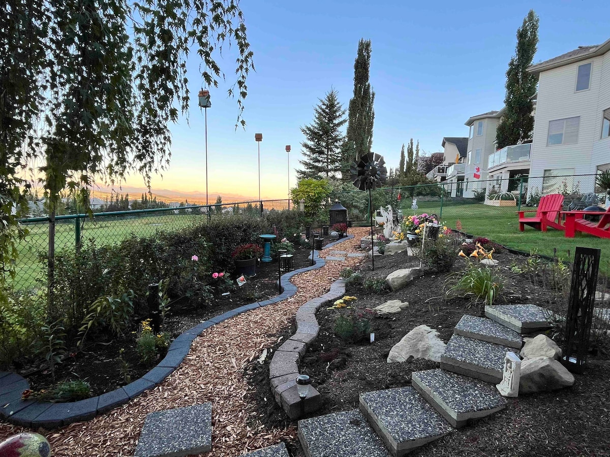 A landscaped garden path winds through colorful flowers and plants, bordered by elegant stone steps. Subtle lighting accents the pathway, leading to a serene sitting area with distinct views of the rolling landscape. A picturesque sunset casts a warm glow over the tranquil scene.