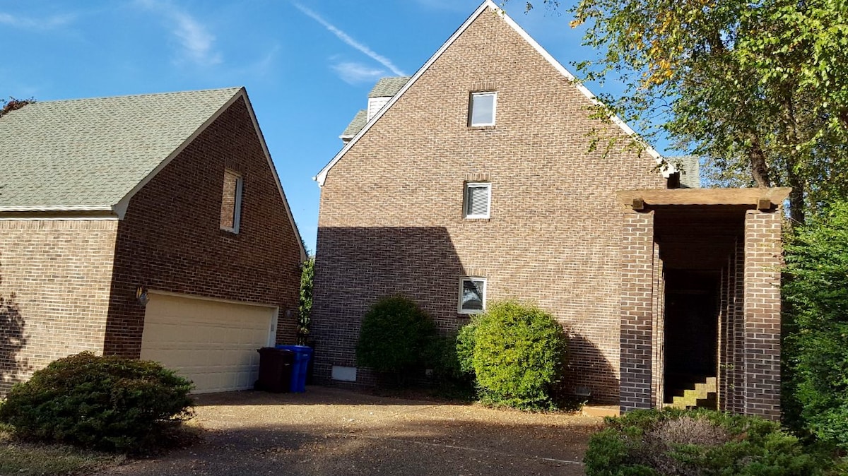 The exterior of a two-story brick apartment building is shown, featuring a staircase leading to the entrance. Lush green shrubs and trees frame the entrance, while a driveway accommodates two blue recycling bins. A neighboring structure is visible to the left.