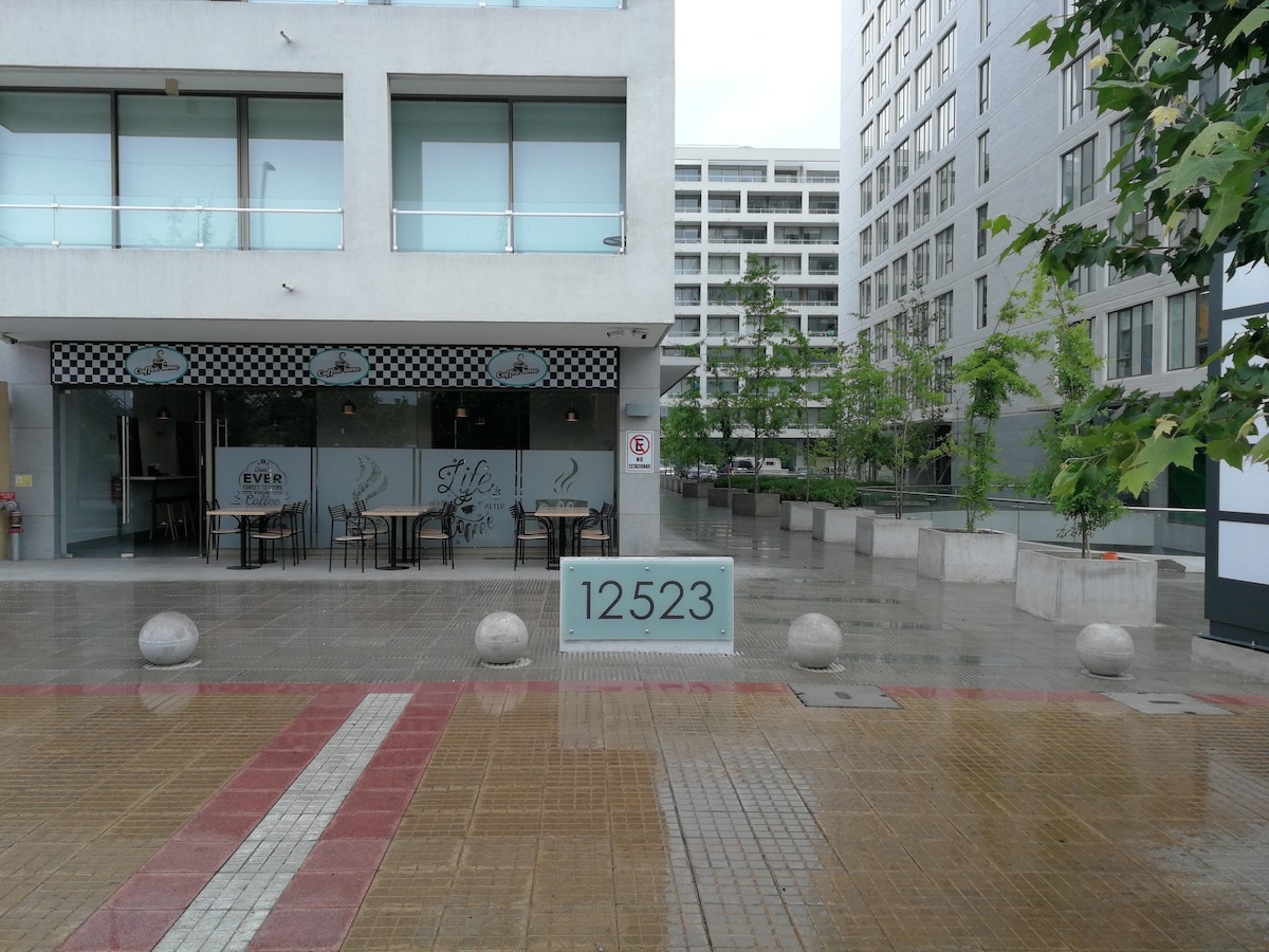 A modern building facade is seen featuring large glass windows and a restaurant entrance. A number '12523' is displayed prominently in front. The pavement is made of tiles, with stone planters and trees enhancing the outdoor environment.