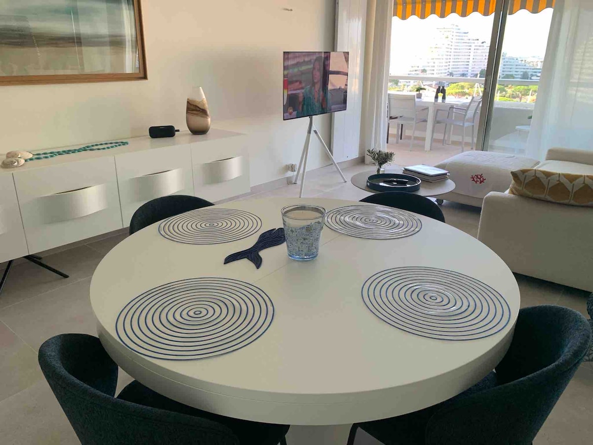 A round dining table is centered within the room, surrounded by six black chairs. Simple placemats design the table surface. A television is mounted on a stand nearby, with a glass vase positioned on the table. Natural light enhances the inviting atmosphere.