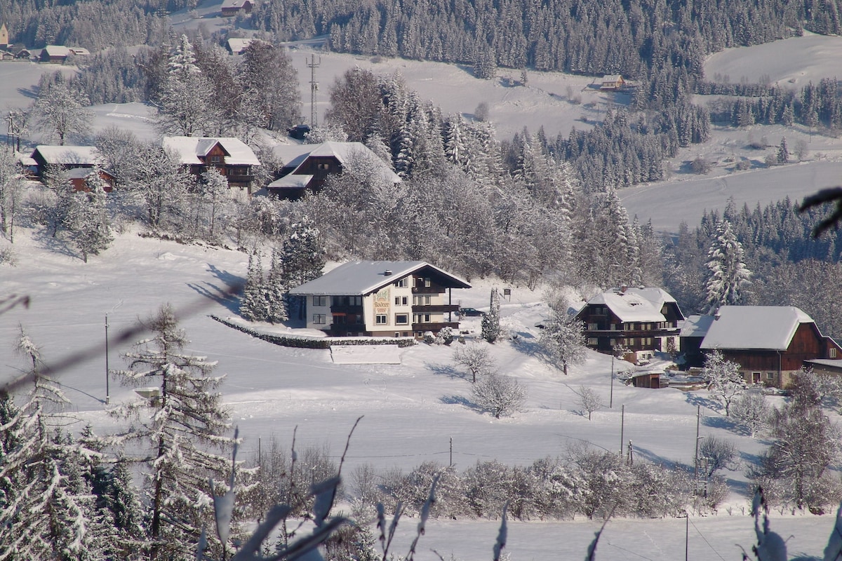 A serene winter landscape is depicted with a cluster of charming chalets blanketed in fresh snow. The surroundings feature snow-covered trees and distant hills, contributing to a peaceful alpine atmosphere. The primary house is prominent with its modern architectural style, surrounded by traditional wooden structures.