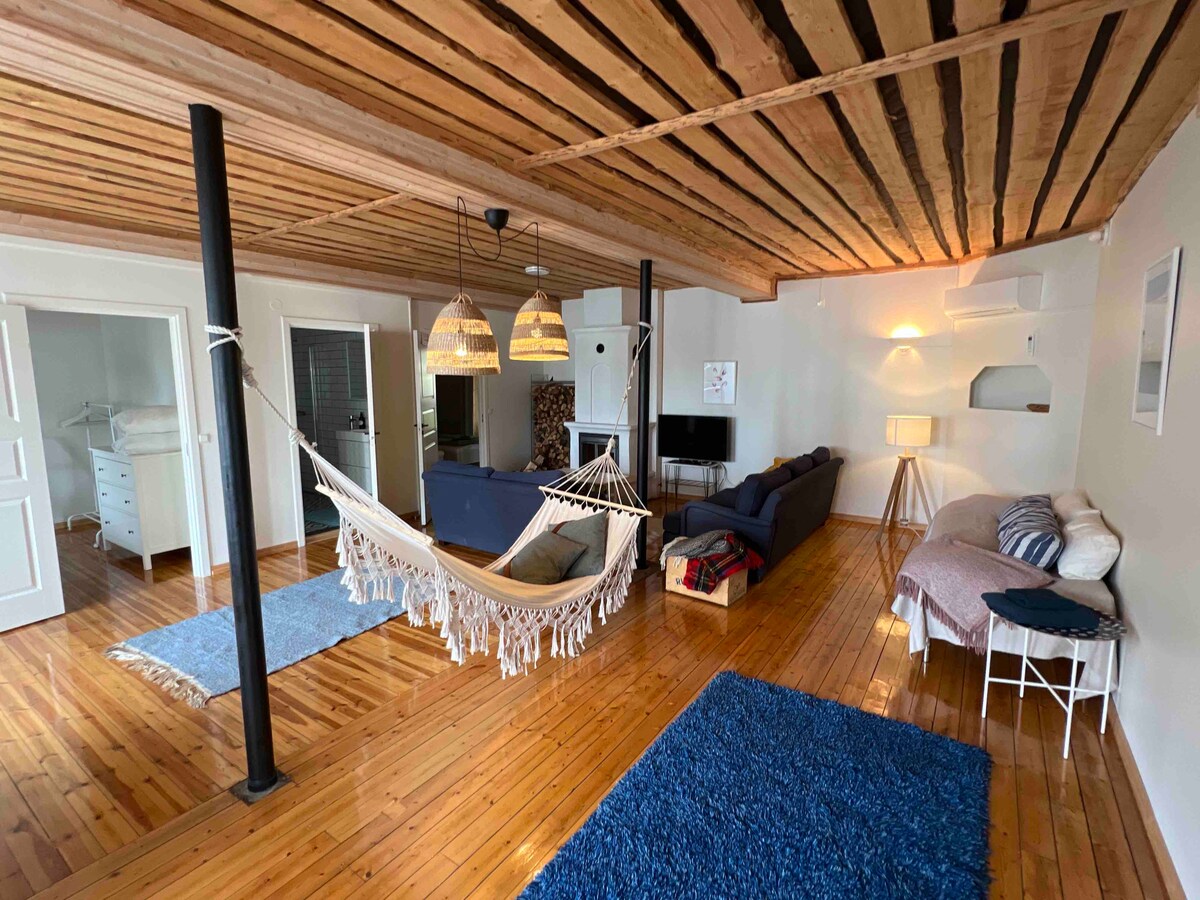 A spacious living area featuring warm wooden floors and a cozy hammock. Soft furnishings include a dark blue sofa and a light-colored bed. The ceiling showcases rustic wooden beams, while pendant lights provide gentle illumination. A television is positioned in the corner, and textured rugs add comfort.