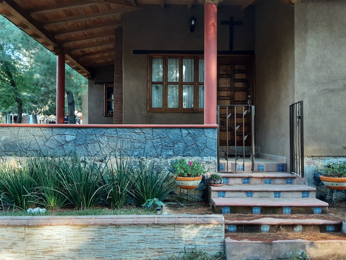 A welcoming entrance features a set of stone steps leading to a sturdy wooden door. Flower pots are arranged along the pathway, and a cross is mounted above the entrance. Lush greenery enhances the tranquil atmosphere of the front yard.