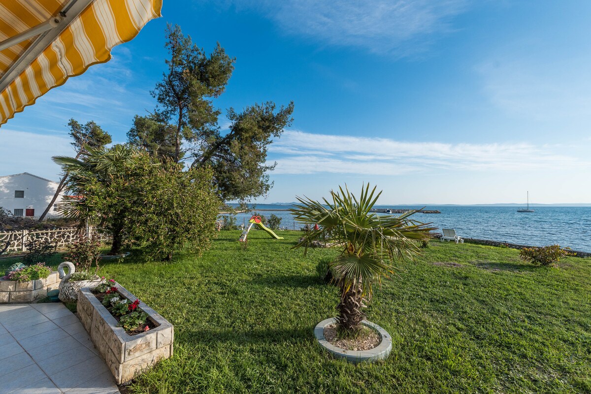 A well-maintained garden area features lush green grass and tropical plants, including a palm tree. A clear view of the sea is visible in the background, with a few distant boats and a blue sky above. Comfortable seating options can be seen along the edge of the garden.