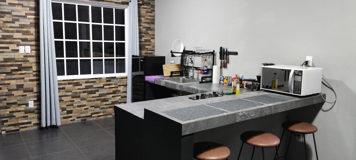A modern kitchen area is presented with a spacious countertop and black bar stools. A stove is positioned on the counter, complemented by a microwave and various kitchen utensils. Natural light enters through a large window, accentuated by light grey curtains, against a backdrop of textured wall tiles.