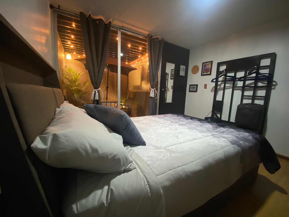 A spacious sleeping area features a large bed with crisp white linens and decorative pillows. Natural light filters in through floor-to-ceiling windows adorned with dark curtains, revealing a glimpse of the outdoor terrace area with twinkling lights.