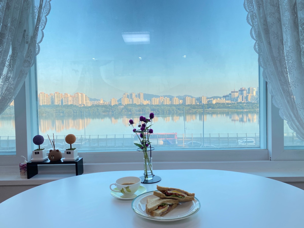 A table is set with a plate of sandwiches and a cup of tea, positioned near a window that offers a stunning view of the Han River and the city skyline. Soft curtains frame the scene, while a small vase with colorful flowers adds a touch of elegance.