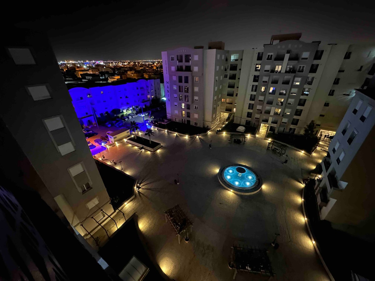 An aerial view of an illuminated courtyard at night, showcasing a central circular fountain surrounded by modern residential buildings. Soft blue lighting highlights the area, while pathways are lined with light, enhancing the outdoor space. The scene captures a serene ambiance in a contemporary setting.