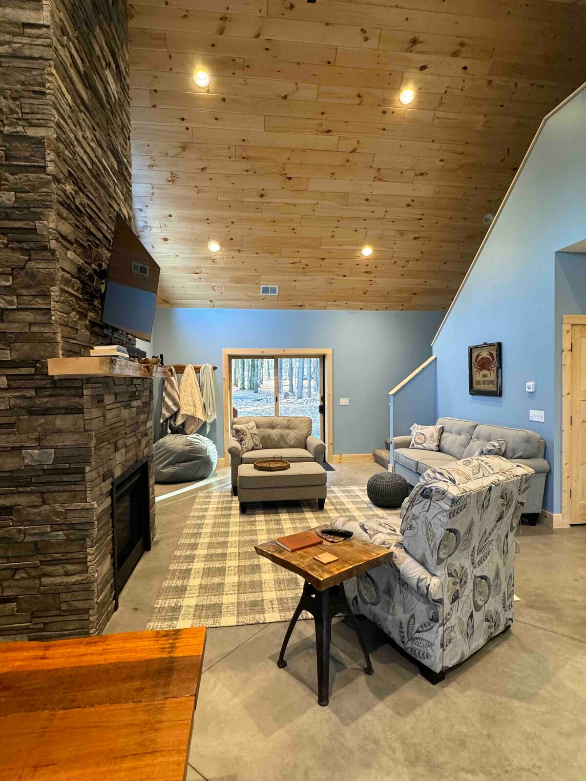An inviting living area features a stone fireplace framed by wooden paneling. Soft seating arrangements, including a light blue sofa and patterned chairs, are positioned around a central coffee table. Large windows allow natural light to fill the space, revealing views of the surrounding woods.
