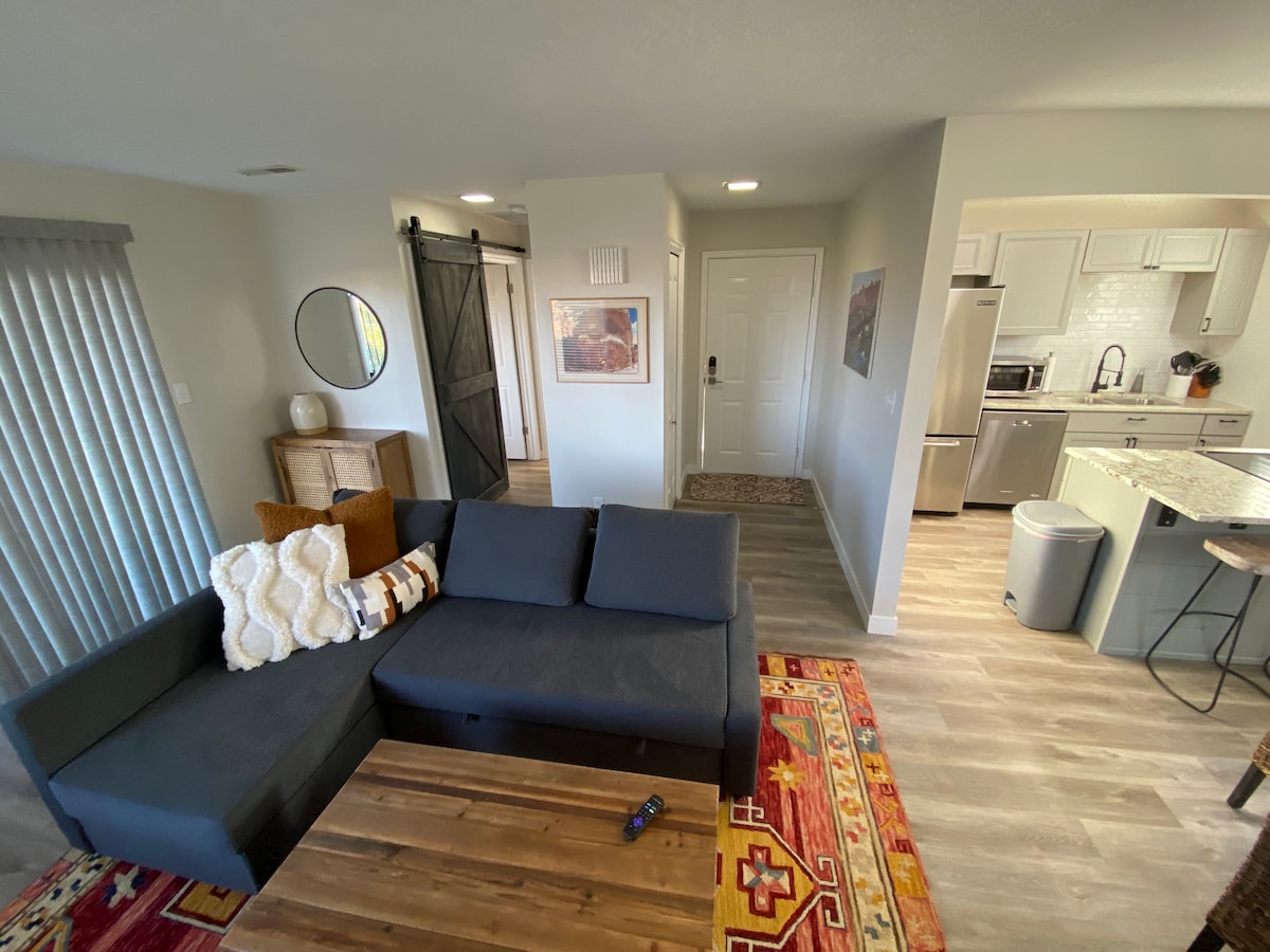 A cozy living area features a dark sectional sofa paired with decorative pillows. A wooden coffee table is positioned on a colorful area rug. The space includes a mirror and a sliding barn door, leading to a welcoming entrance and a well-equipped kitchen.