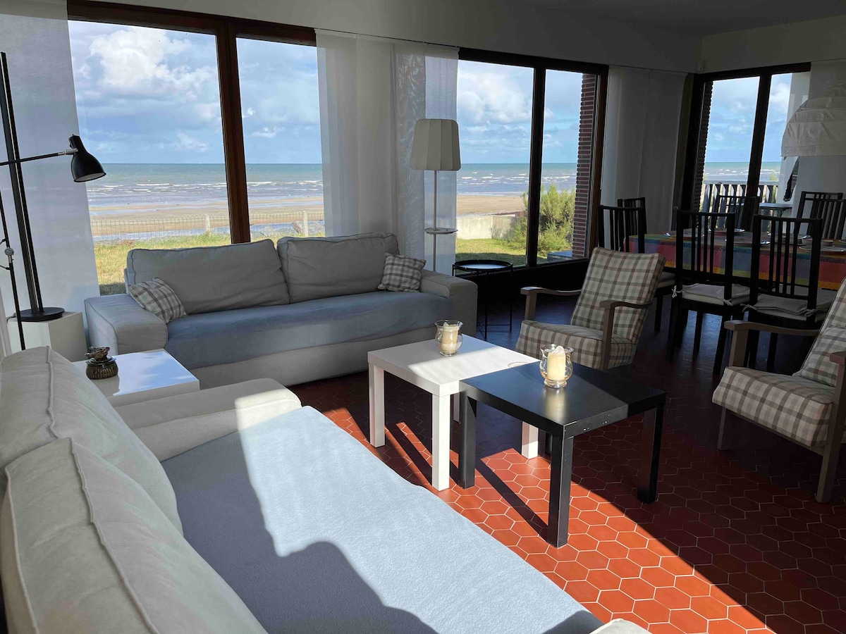 A spacious living area features large windows overlooking the sea, allowing natural light to fill the room. Comfortable sofas are arranged around a low table, complemented by a dining area with chairs in view. The warm colors of the flooring add a cozy touch.