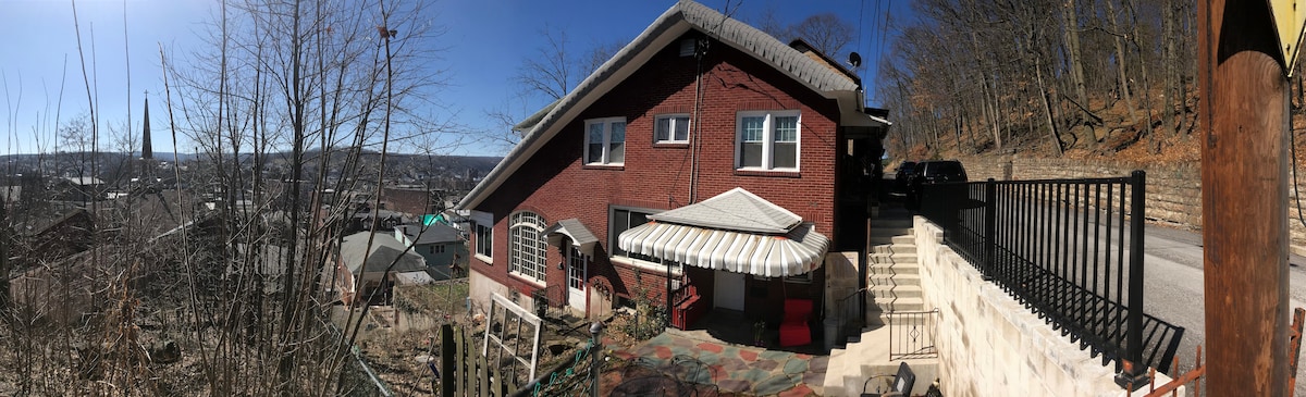 The exterior of a red brick apartment is displayed, showcasing a sloped structure nestled on a mountainside. The entrance features an awning, with stone steps leading up to the entrance. Surrounding vegetation and a clear sky complete the atmosphere.
