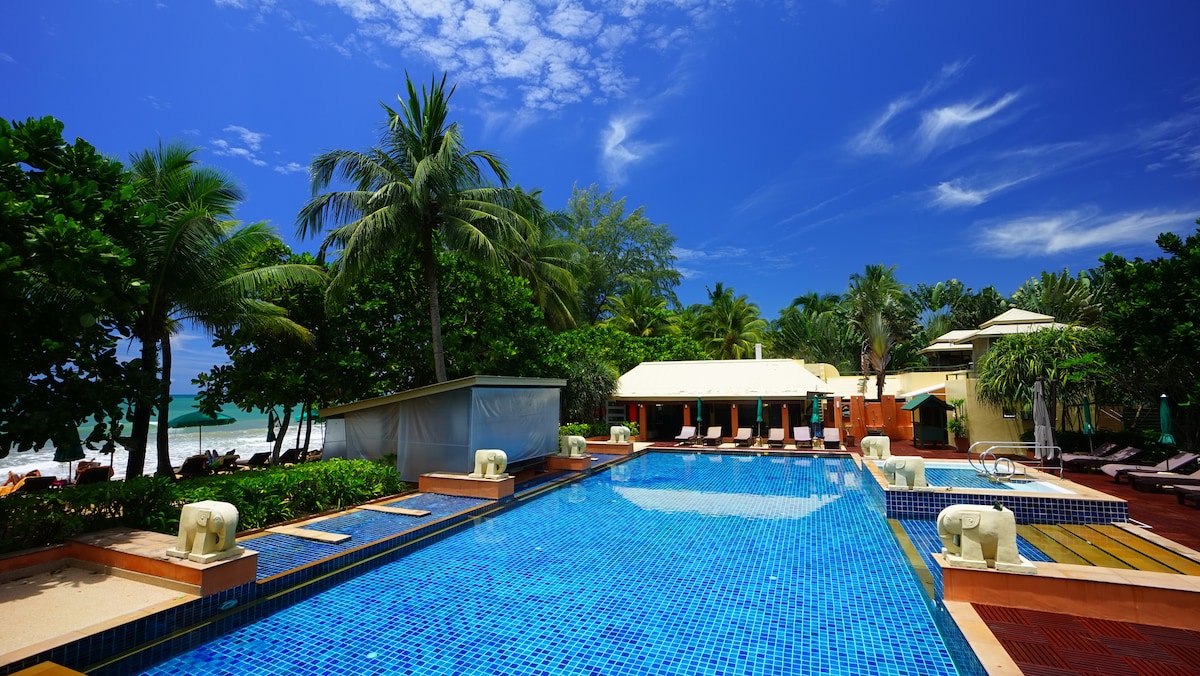 A tranquil pool area is surrounded by lush tropical vegetation, featuring a large blue-tiled pool reflecting clear skies. Sun loungers are arranged nearby, and a white building with a sloped roof is situated in the background, enhancing the serene environment.