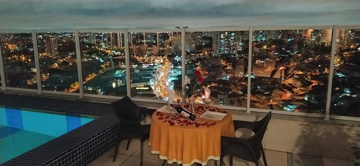 A dining table is set with a yellow tablecloth on a balcony overlooking a city skyline at night. The table is adorned with romantic elements, including rose petals and a decorative bottle. A sparkling pool lies nearby, reflecting the city lights.