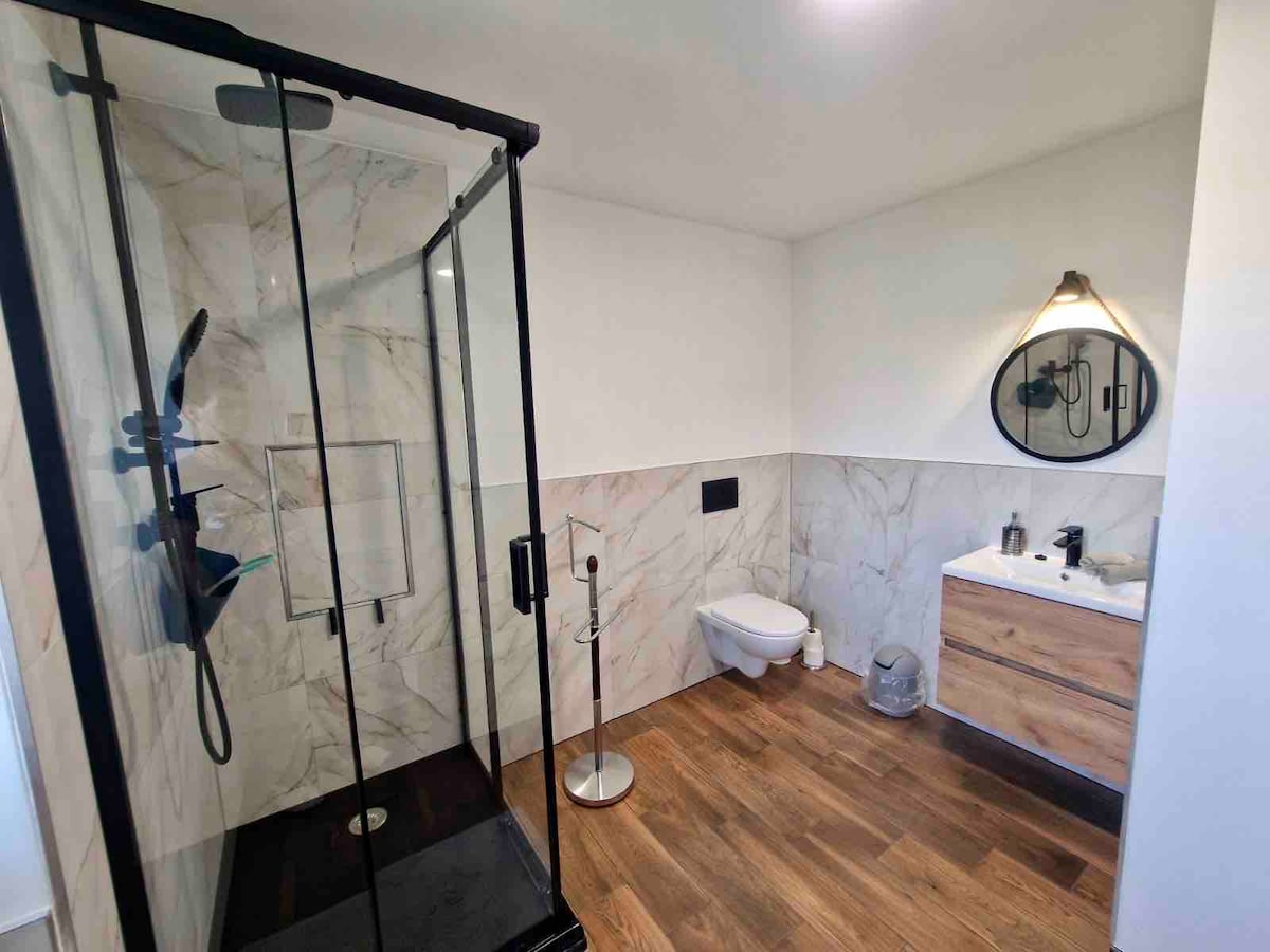 A modern bathroom features a glass-enclosed shower with marble walls and a rainfall showerhead. A wooden vanity with a round mirror is visible, accompanied by a sleek toilet. Natural light brightens the space, highlighting the warm wooden flooring.