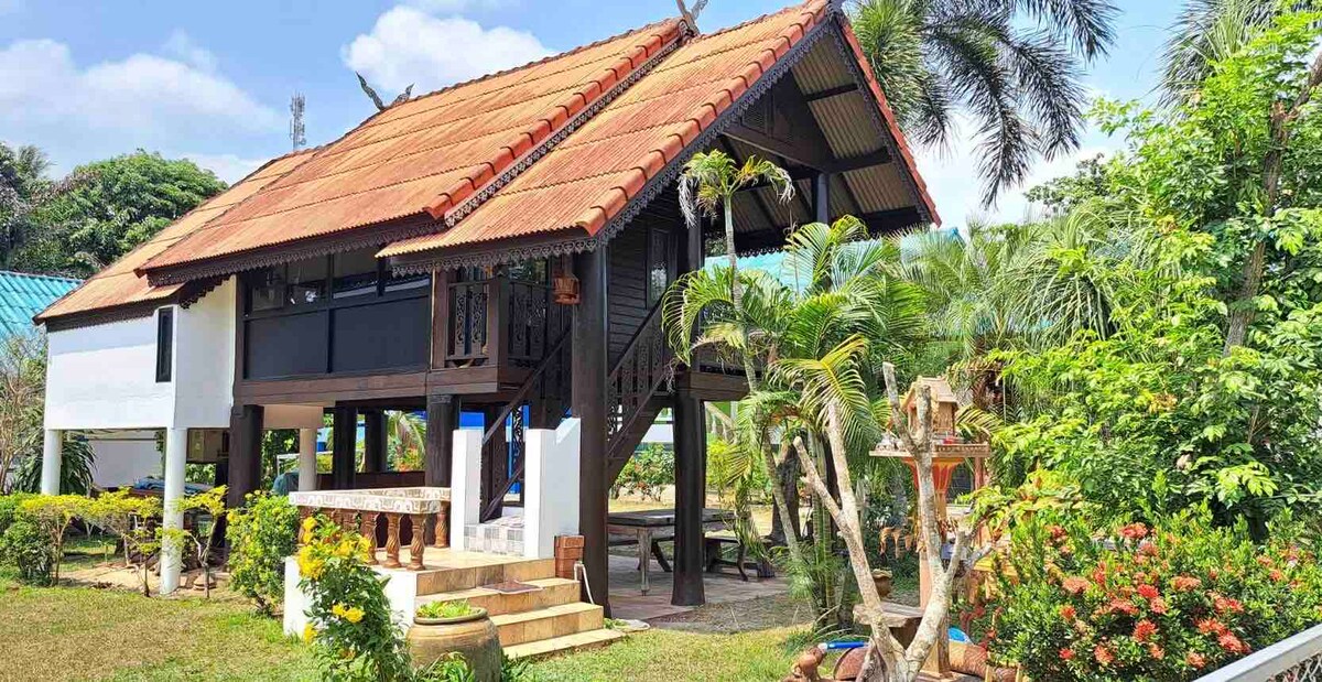 A traditional bungalow-style house is surrounded by lush greenery and colorful flowering plants. The distinct thatched roof overhangs a spacious outdoor area featuring steps leading to the entrance. Towering palm trees frame the property, contributing to a serene and natural atmosphere.
