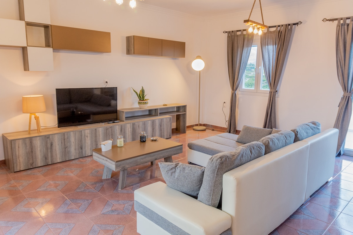 A spacious living area features a modern sectional sofa with gray and white upholstery, complemented by a wooden coffee table. A large television is mounted on the wall above a sleek console. Natural light enters through a window draped with sheer curtains, enhancing the room's relaxed ambiance.