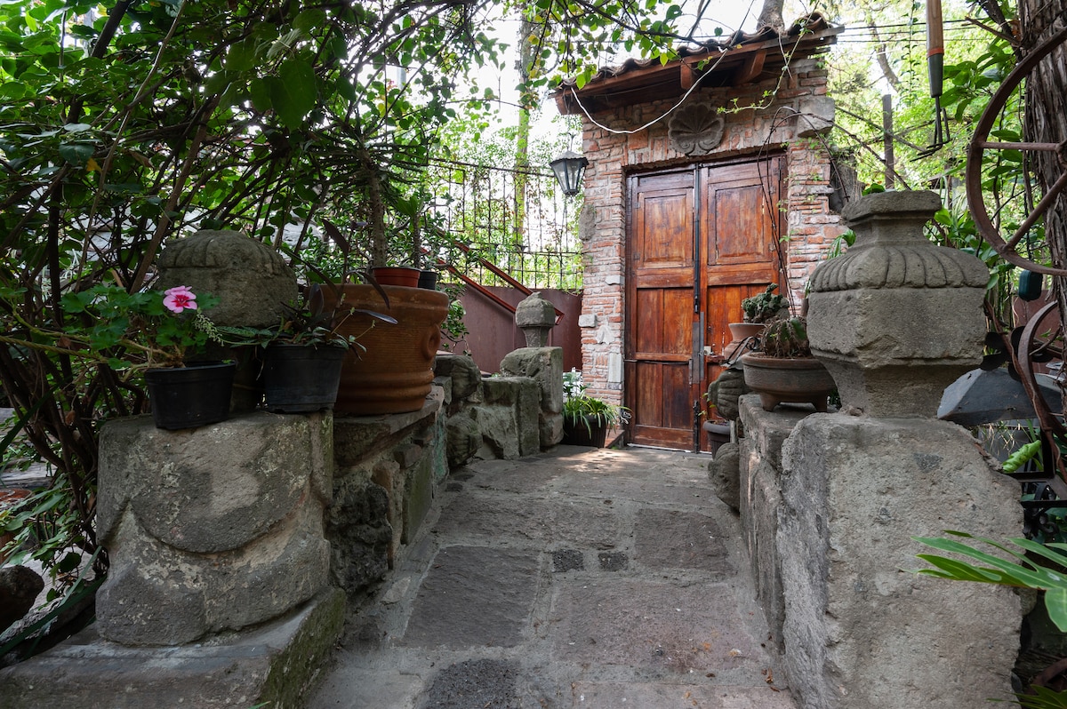 A stone pathway leads to a wooden door framed by lush greenery and various potted plants. The entrance is adorned with rustic stone walls, showcasing the original materials and craftsmanship of the surrounding area. Sunlight filters through the foliage, creating a serene atmosphere.