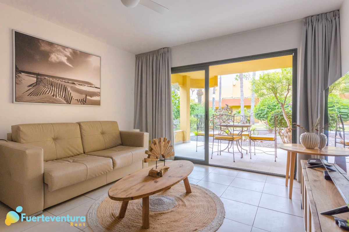 A light-filled living area features a beige sofa paired with a wooden coffee table and a woven rug. Large glass doors open to a patio with outdoor seating, enhancing the view of the tropical garden. Wall art depicting a beach scene adorns one side.