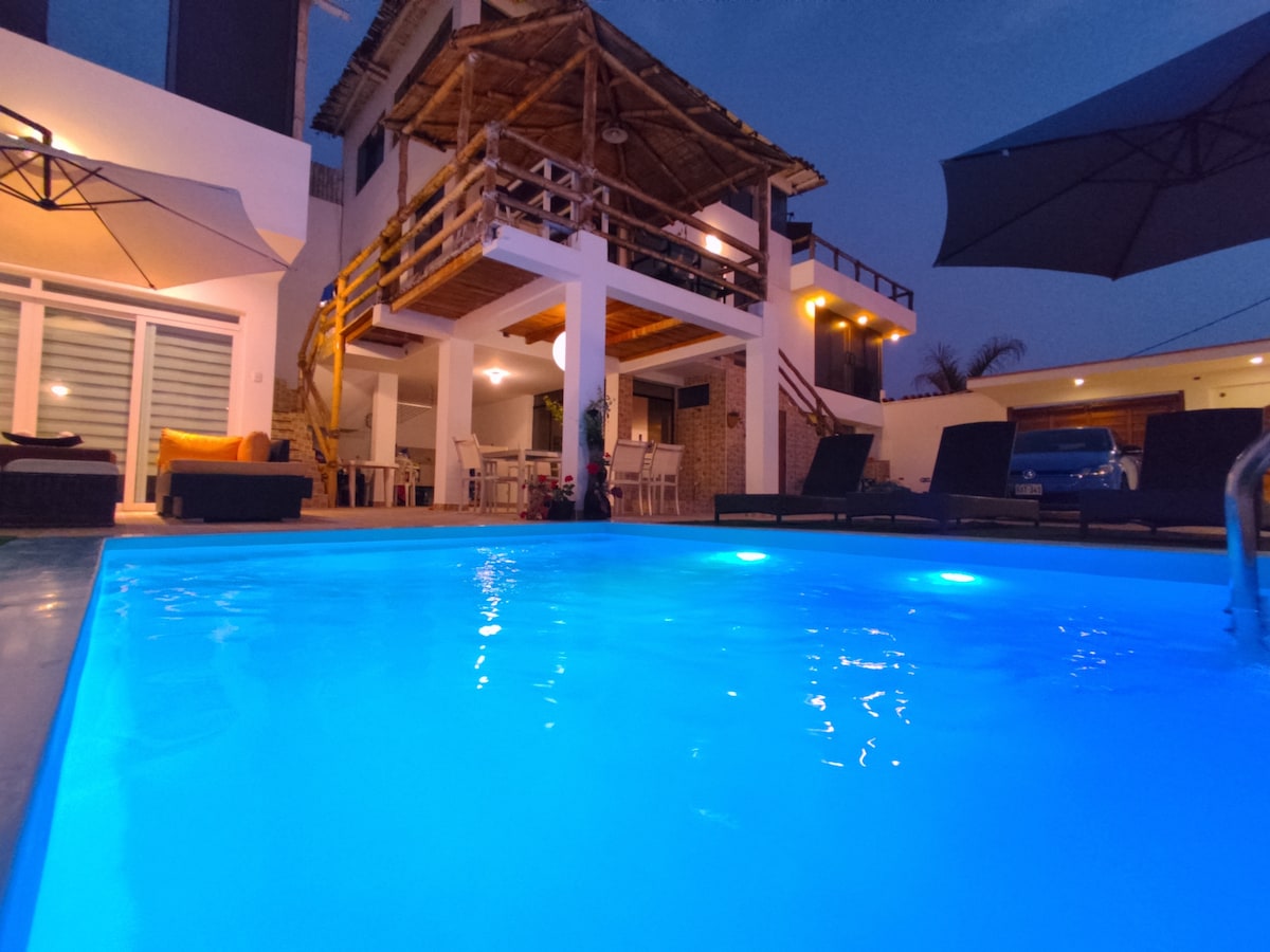 The illuminated pool appears inviting under the evening sky, reflecting soft blue hues. Surrounding the pool, lounge chairs provide a comfortable seating area, while a spacious terrace above offers additional space for relaxation. The architecture features wooden elements, enhancing a natural ambiance.
