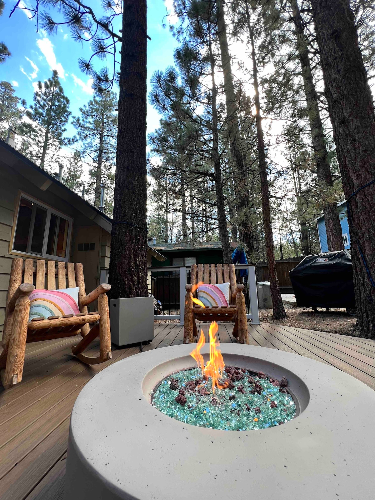 A cozy outdoor space features two rustic rocking chairs positioned near a round fire pit with colorful glass stones and an active flame. Pine trees surround the area, and a propane barbecue is visible in the background.