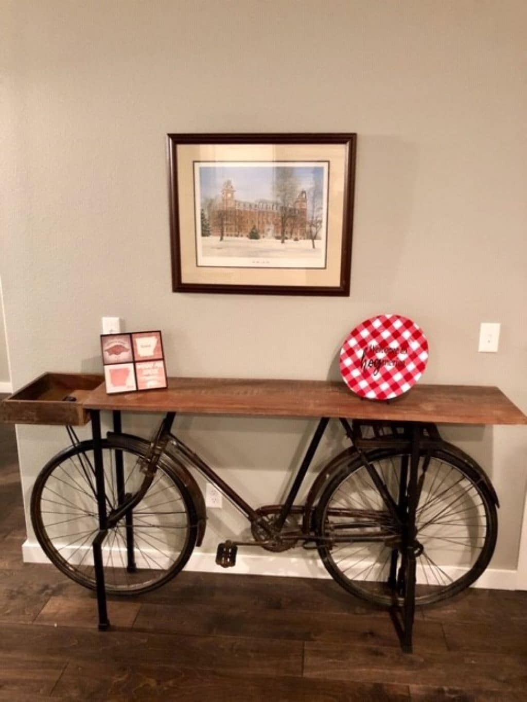 A rustic wooden console table is supported by the front wheel of a vintage bicycle, positioned against a light gray wall. Above, a framed artwork displays a historic building, while decorative items, including a red and white checked plate and framed photos, are arranged on the table.