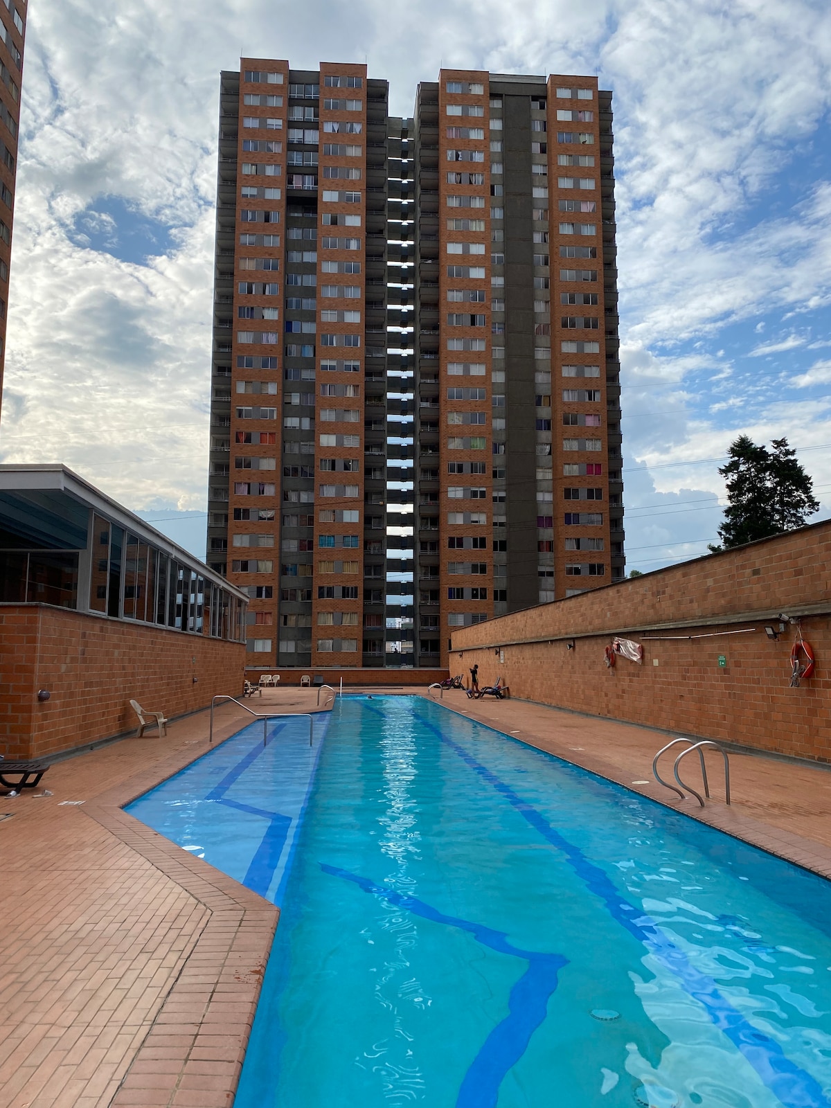The image depicts a long swimming pool enclosed by brick walls. Adjacent buildings rise prominently in the background, with multiple stories visible. The sky features light clouds, contributing to the overall open air of the environment.