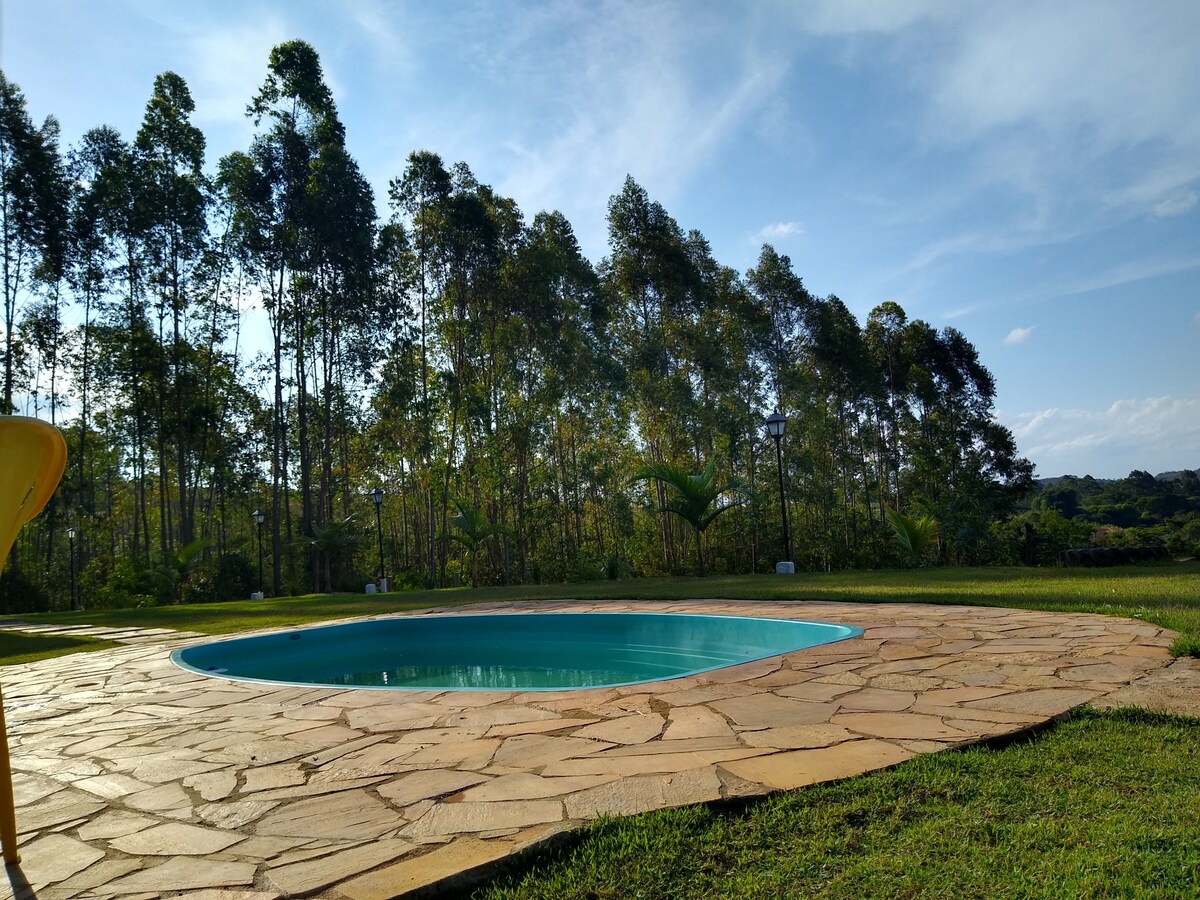 A refreshing swimming pool is set against a backdrop of tall, lush trees under a clear blue sky. The pool area features a natural stone patio, surrounded by green grass, providing a serene environment ideal for relaxation.
