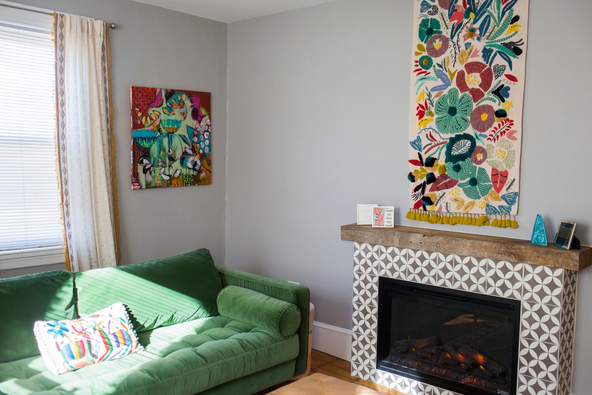 A vibrant green couch is positioned next to a decorative fireplace. A colorful textile art piece adorns the wall above the fireplace, complemented by another artwork on the adjacent wall. Natural light enters through the window, illuminating the space lightly.