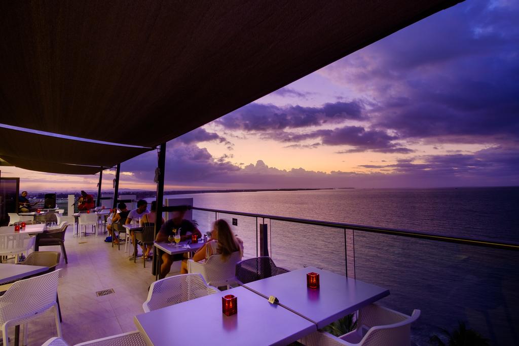 An outdoor dining area features modern tables and chairs, arranged to face the ocean. Soft lighting is provided by candles on the tables, while a large canopy offers shade. A vibrant sunset casts hues of purple and orange over the water and sky.
