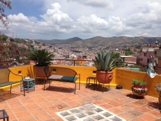 A private terrace is featured with terracotta tiles and surrounded by vibrant plants. Two seating areas are arranged for relaxation, offering panoramic views of the cityscape and mountains under a partly cloudy sky.