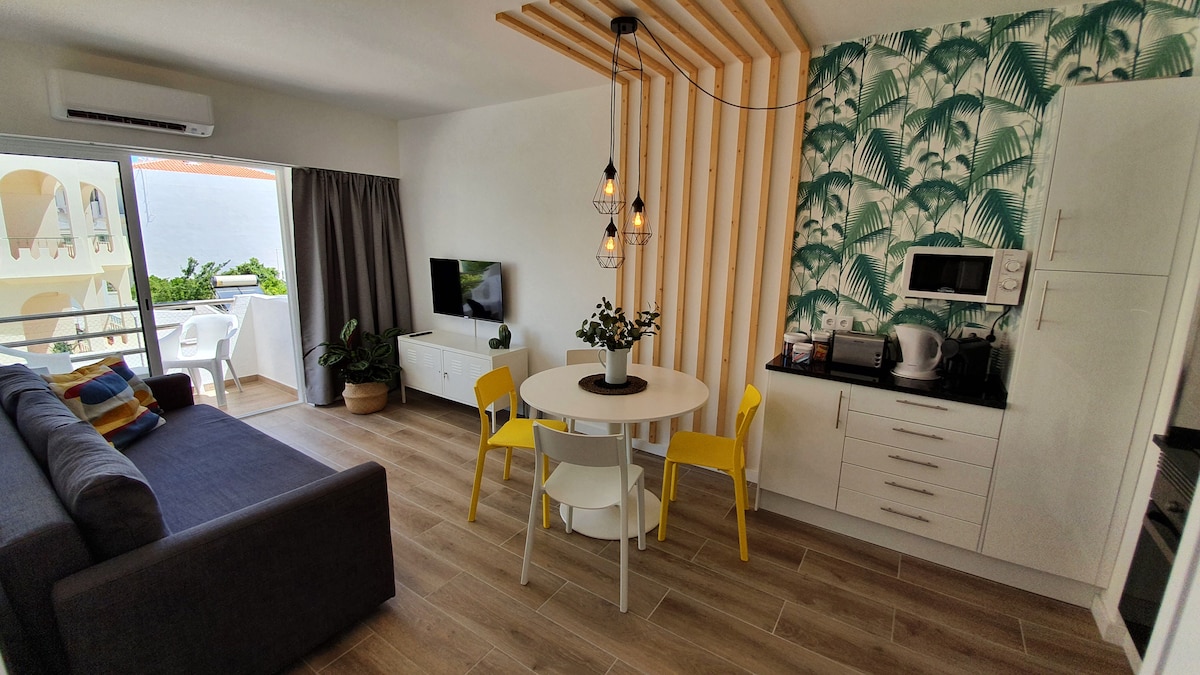 A modern living area features a cozy gray sofa and a round dining table surrounded by yellow chairs. A wall adorned with green tropical patterns adds a lively touch. A flat-screen TV is positioned near large windows, allowing natural light to enter.