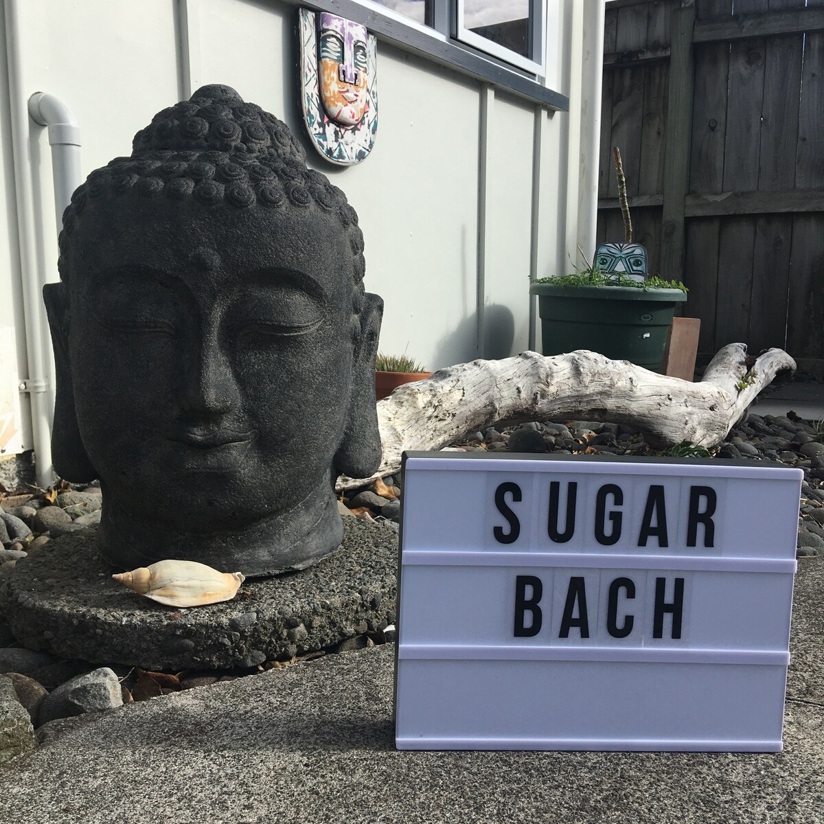 A stone Buddha statue is placed on a textured surface in the foreground, accompanied by a decorative shell. A sign reading 'SUGAR BACH' is positioned nearby. The background features a wooden fence and various plants, creating an inviting outdoor ambiance.