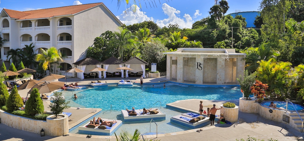 A large pool area is surrounded by lush tropical landscaping. Several loungers and shaded cabanas are positioned around the pool. Guests are seen enjoying the spacious water features, while palm trees provide a relaxed ambiance against a clear blue sky.