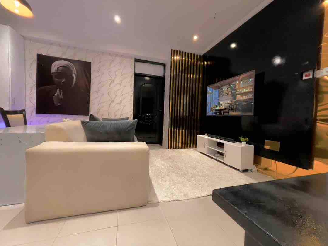 A modern living area features a light-colored sofa, complemented by decorative cushions. A plush rug covers the tiled floor, while an entertainment unit supporting a flat-screen television is displayed against a dark accent wall. Ambient lighting creates a relaxed atmosphere.