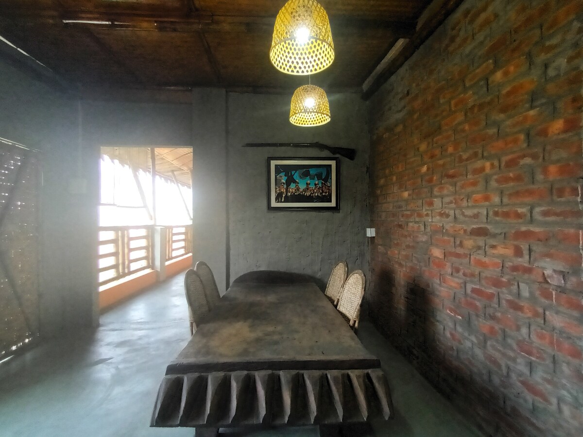A spacious dining area is captured, featuring a large wooden table surrounded by woven chairs. Natural light filters through a nearby open space with a view of a balcony. Two woven pendant lights hang from the ceiling above the table, enhancing the inviting atmosphere.