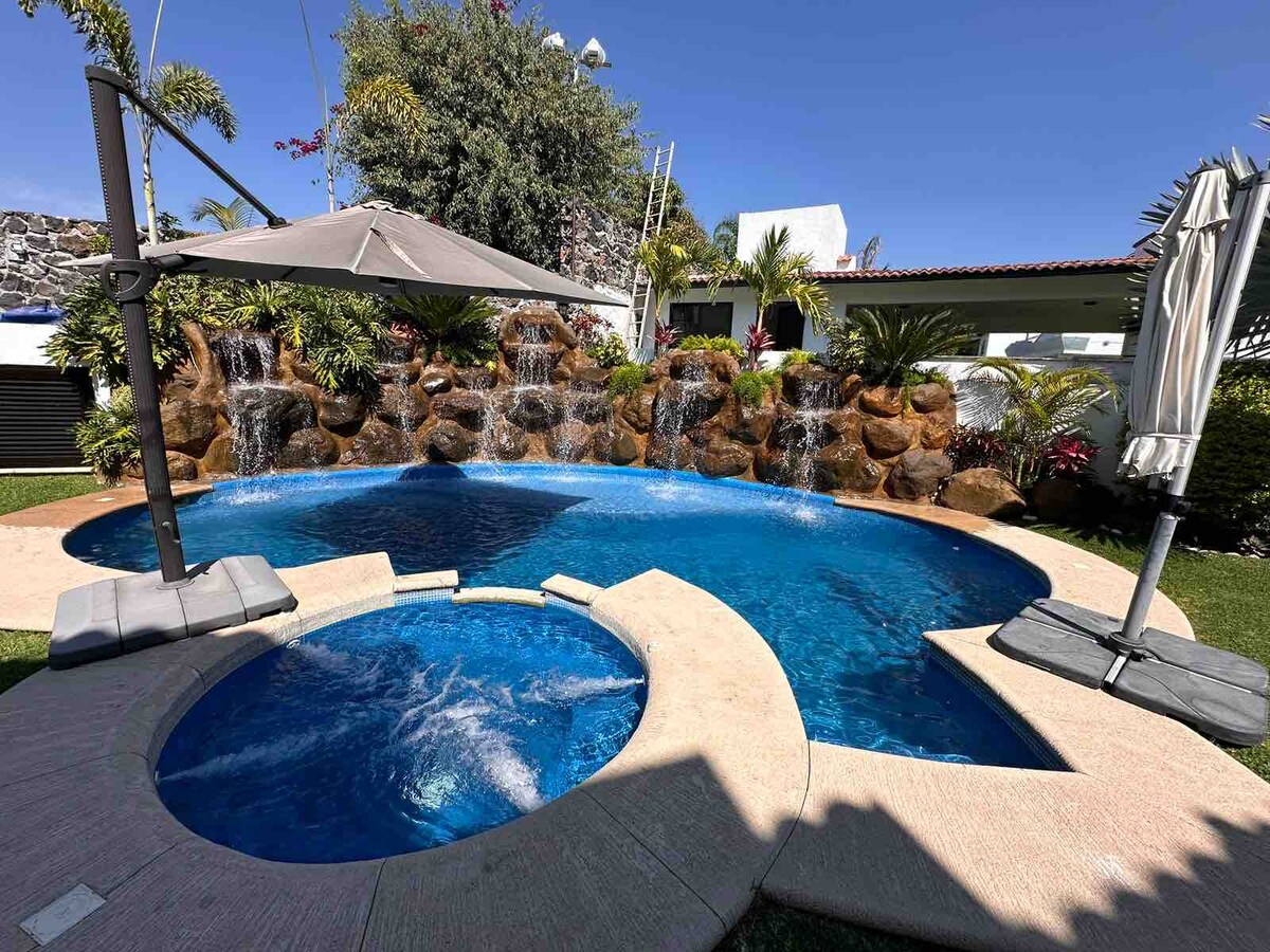 A refreshing outdoor area features a large pool with a jacuzzi attached, surrounded by a stone waterfall that adds a tranquil sound. Sun loungers with shades are positioned beside the water, complemented by lush greenery around the space.