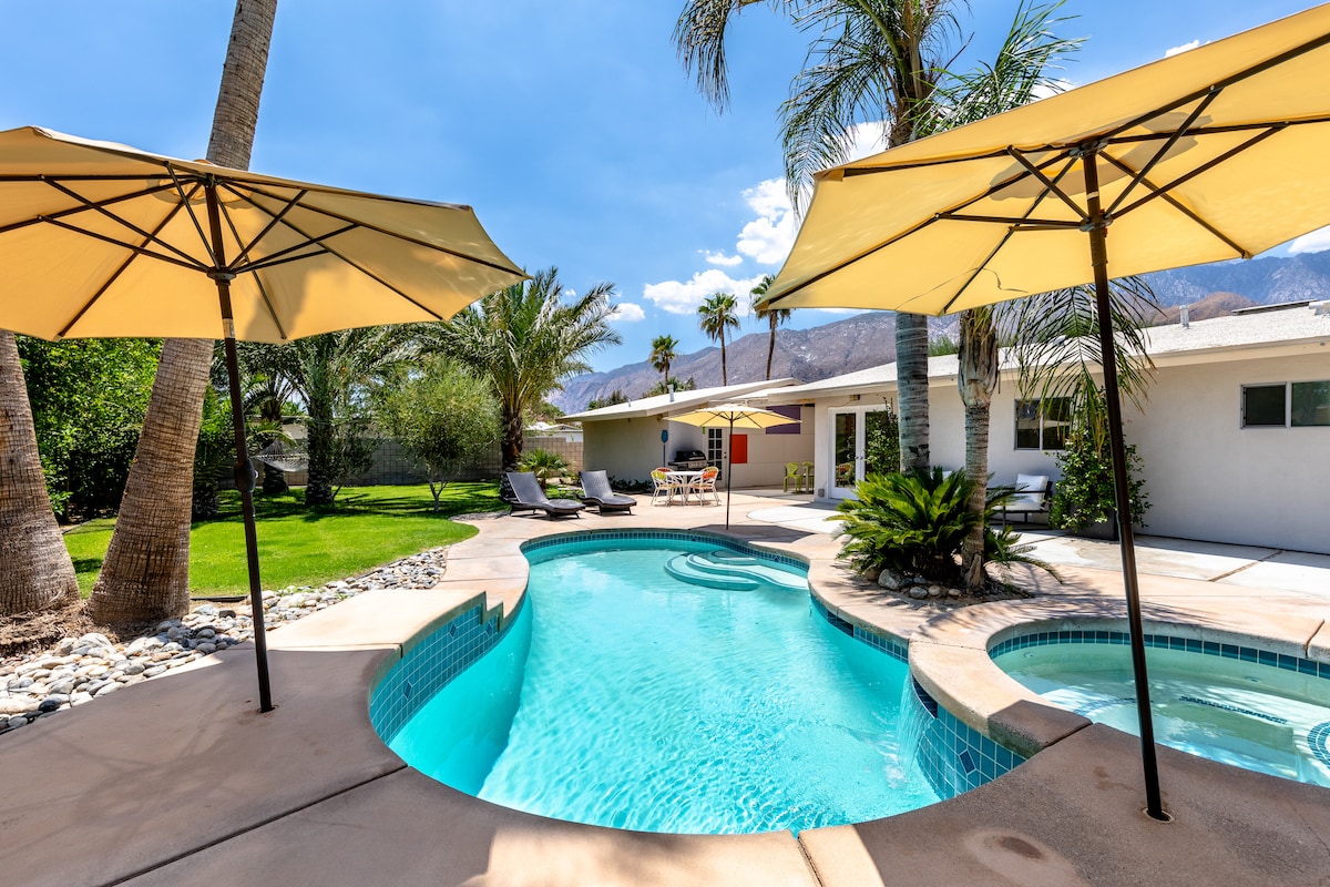 A spacious salt-water pool is framed by inviting lounge chairs and yellow umbrellas, providing shade. Lush palm trees surround the area, enhancing the outdoor oasis. The jacuzzi is integrated alongside the pool, with a view of the mountains in the background.