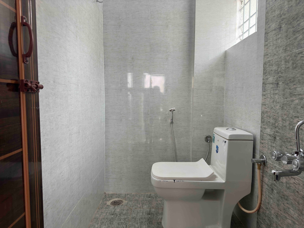 A modern bathroom features smooth, gray stone walls and floor tiles. A toilet is positioned alongside a sleek shower area, equipped with a handheld shower head. Natural light filters in through a small window, enhancing the spacious feel of the room.