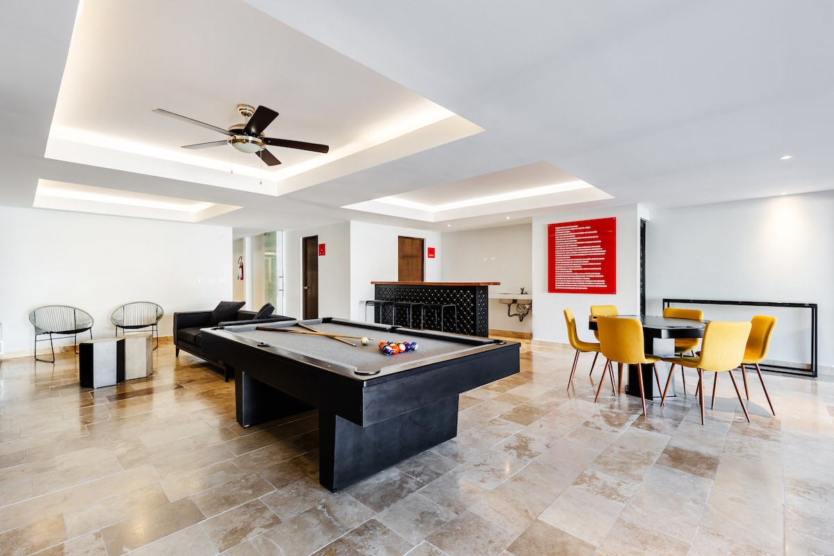 A spacious common area features seating options, including two yellow chairs and modern black accents. A sleek pool table is centered in the room, surrounded by polished stone flooring. Natural light fills the area, accentuated by a ceiling fan and ambient lighting along the ceiling.