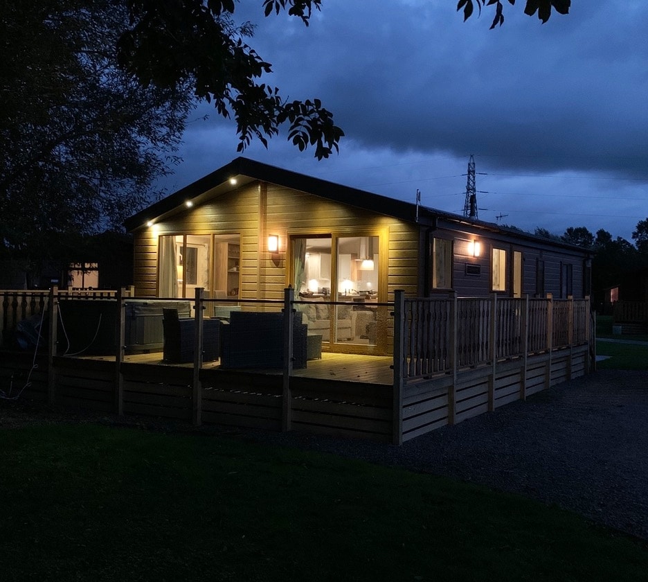 The lodge is illuminated against the evening sky, showcasing a spacious deck surrounded by a wooden railing. Bright lights highlight the exterior, with large windows offering a view into the welcoming interior. Comfortable outdoor seating invites relaxation and enjoyment of the surroundings.