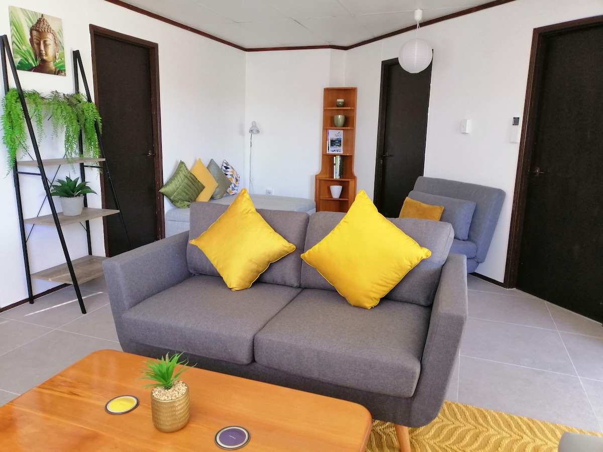 A cozy living area features a gray sofa adorned with vibrant yellow cushions. A wooden coffee table sits in front, complemented by a decorative plant. A small bookshelf and a comfortable chair complete the space, illuminated by natural light streaming through nearby windows.