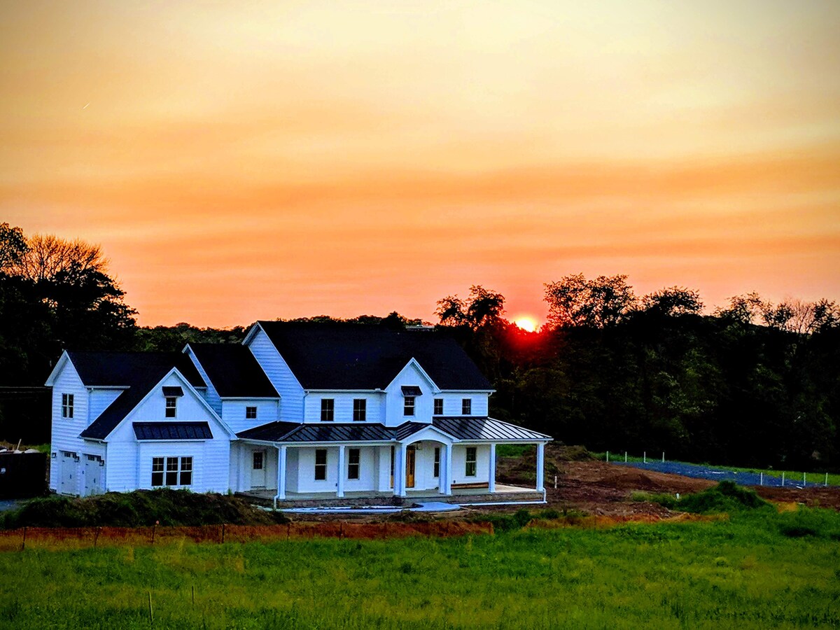 A modern white house is showcased against an orange and pink sunset sky, with the sun partially visible on the horizon. The home's covered porch and multiple windows create an inviting appearance, surrounded by green grass and a natural landscape.